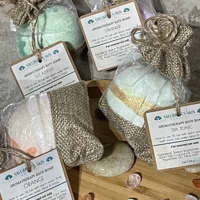 Four aromatherapy bath bombs in burlap bags, each with a different color of liquid inside, and labeled with different scents such as Grapefruit Tangerine and Orange.
